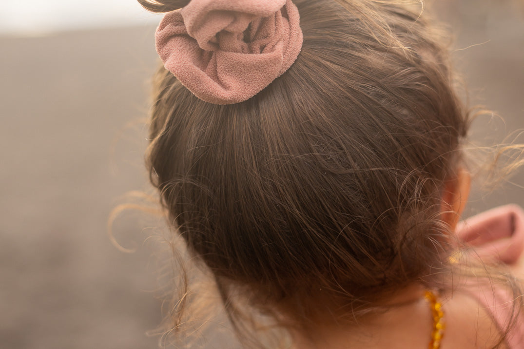 Ein Kind mit braunem Haar zu einem Dutt gebunden, in ein rosa Haargummi gewickelt, einem rosa Oberteil mit gebundenen Schulterträgern und einer Bernsteinkette bekleidet, blickt auf eine unscharfe Strandkulisse.