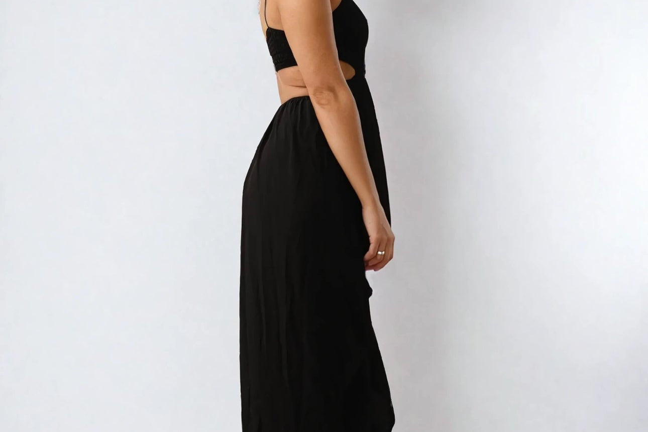 Person in einem schwarzen ärmellosen Top und einem langen Rock steht barfuß seitlich vor einem einfarbigen Hintergrund und blickt in die Kamera.