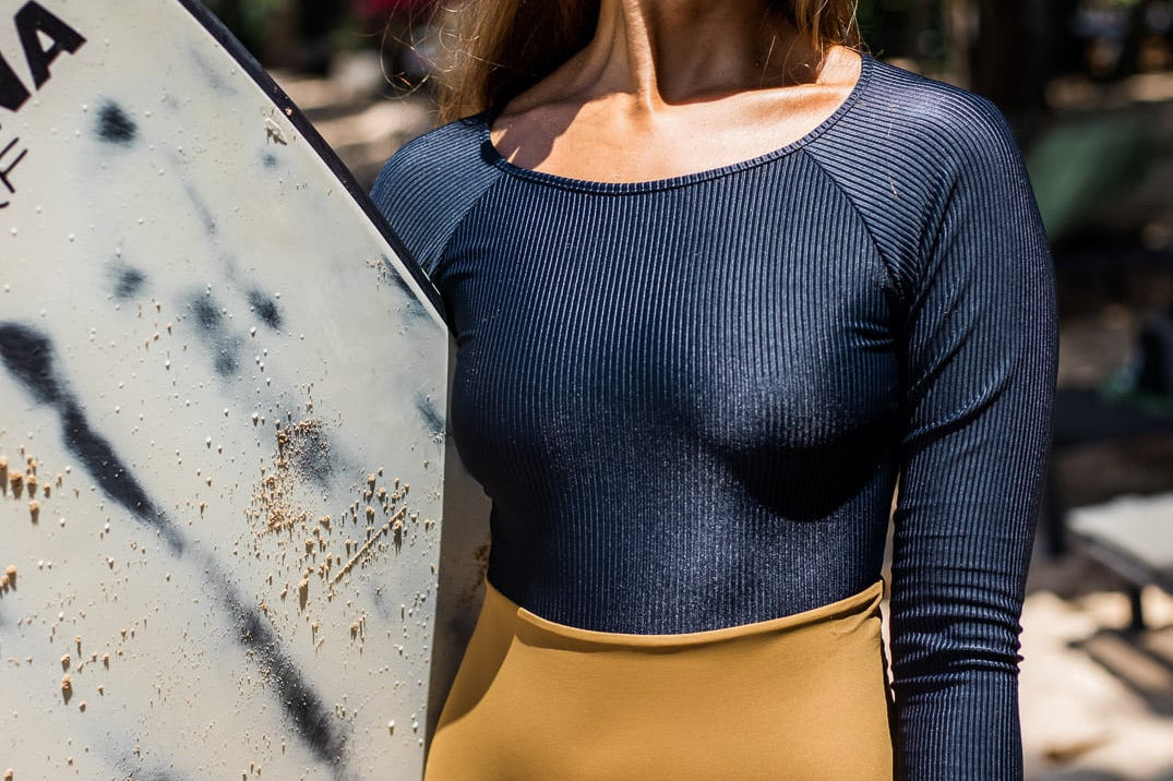 Frau mit Surfbrett an einem sonnigen Strand. Sie trägt einen schwarzen langärmligen Badeanzug und eine hellbraune Hose. Sie hat eine weiße Blume im Haar und blickt zur Seite, im Hintergrund sind Bäume und Strandstühle zu sehen.
