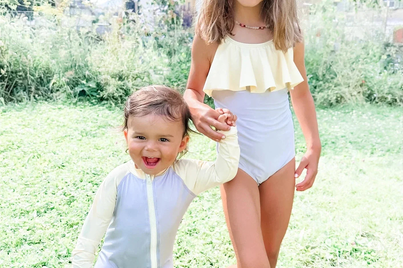 Zwei lächelnde Kinder in Badeanzügen stehen im Gras und halten sich im Freien an den Händen. Das ältere Mädchen trägt einen weiß-gelben gerüschten Badeanzug, das jüngere Kind einen pastellfarbenen langärmeligen Badeanzug.