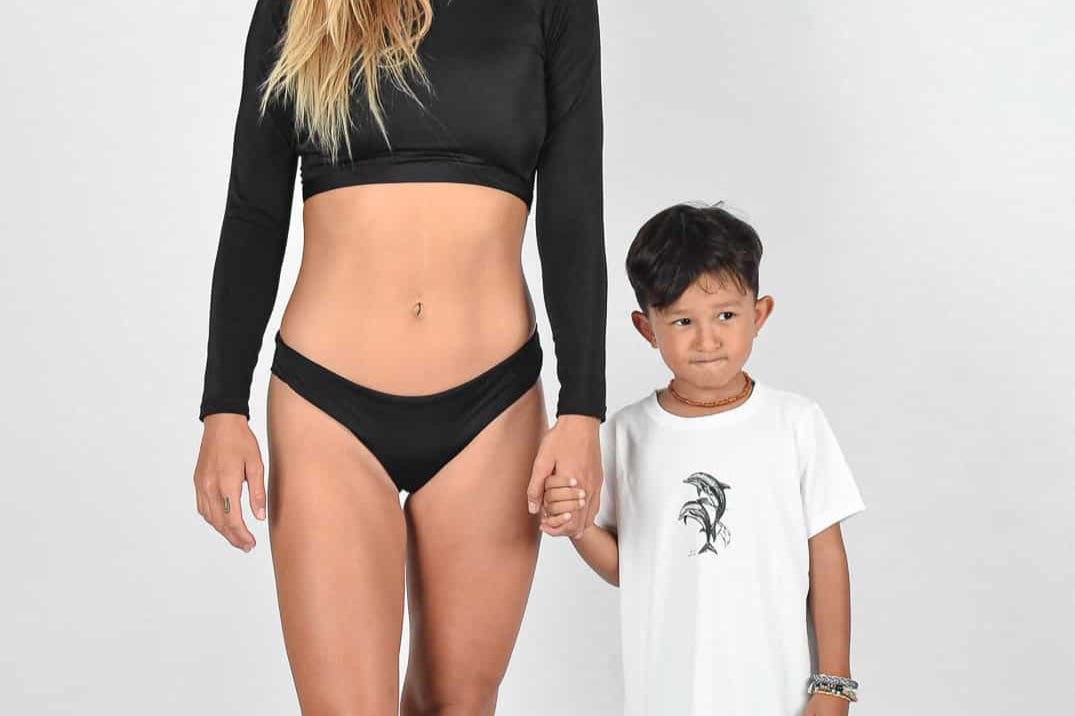 Eine Frau im schwarzen Badeanzug und ein kleiner Junge in weißem Hemd und schwarzen Shorts stehen zusammen vor einem schlichten Hintergrund. Sie lächelt und hält seine Hand.