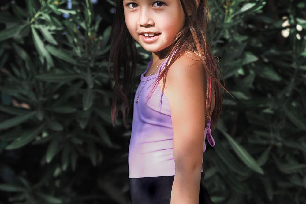 Ein junges Mädchen mit langen braunen Haaren, das einen lila-schwarzen Badeanzug von Main Design trägt, steht im Freien vor grünen Büschen und blickt mit einem leichten Lächeln in die Kamera.