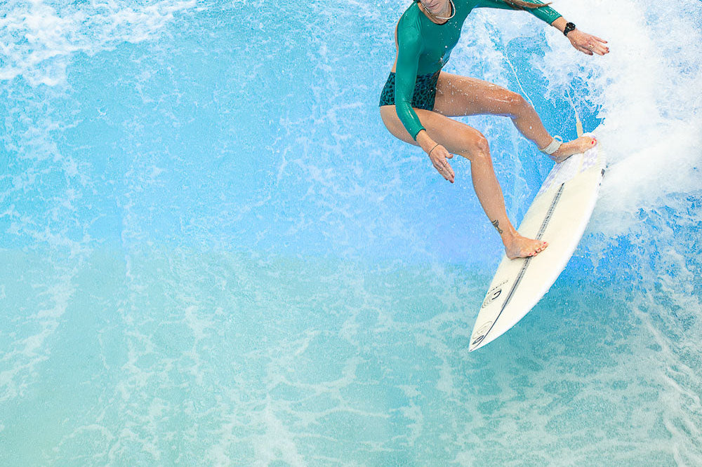 Eine Frau in einem grünen Surfsuit von Main Design reitet auf einem weißen Surfbrett auf einer hellblauen Welle und balanciert geschickt auf dem Wasser, das um sie herum spritzt.