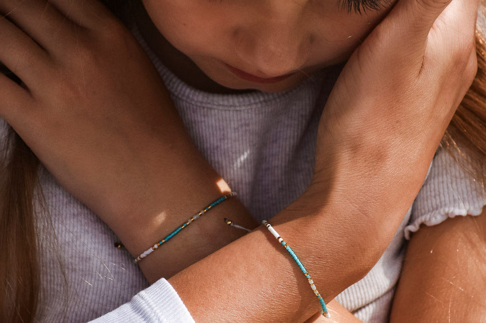 Nahaufnahme eines jungen Mädchens, das sanft von einem Erwachsenen umarmt wird, beide tragen passende zarte Perlenarmbänder. Die Augen des Mädchens sind geschlossen und sie scheint getröstet zu sein.