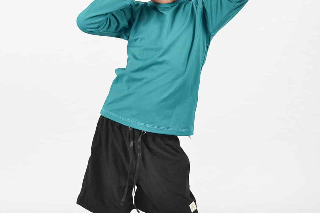 Zwei Kinder in Badeanzügen stehen dicht beieinander am Rande eines Gewässers. Ein Kind hält einen Stock. Nur ihre Rücken und Seiten sind zu sehen. Im Vordergrund sind Gras und Felsen zu sehen.