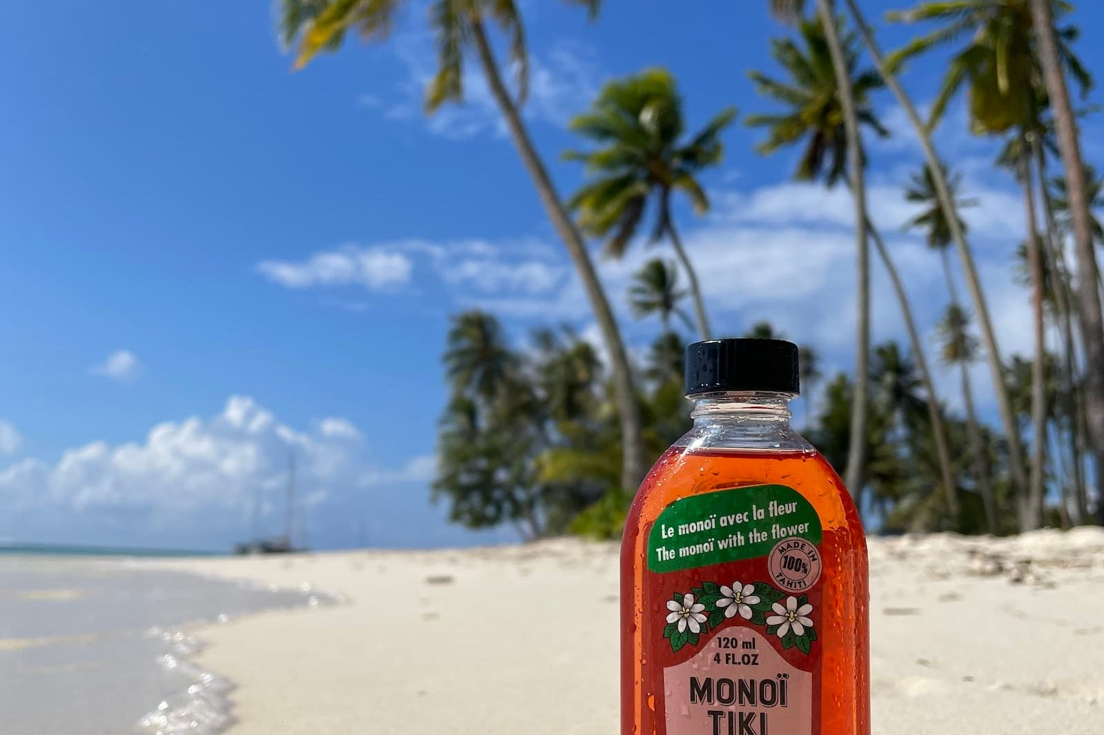 Eine Flasche Monoi Tiki Tahiti Vanille-Bräunungsöl steht an einem Sandstrand mit Palmen und einem klaren blauen Himmel im Hintergrund.