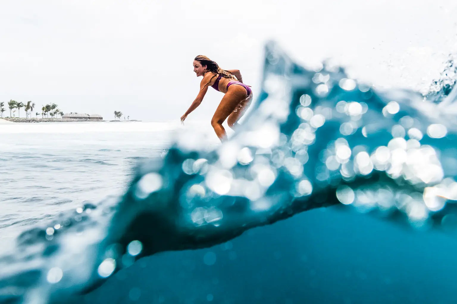 Eine Person surft auf einer Welle im Meer. Der Fokus liegt auf dem klaren blauen Wasser im Vordergrund, während der Surfer und die entfernte Küstenlinie im Hintergrund unter einem bewölkten Himmel leicht verschwommen sind.