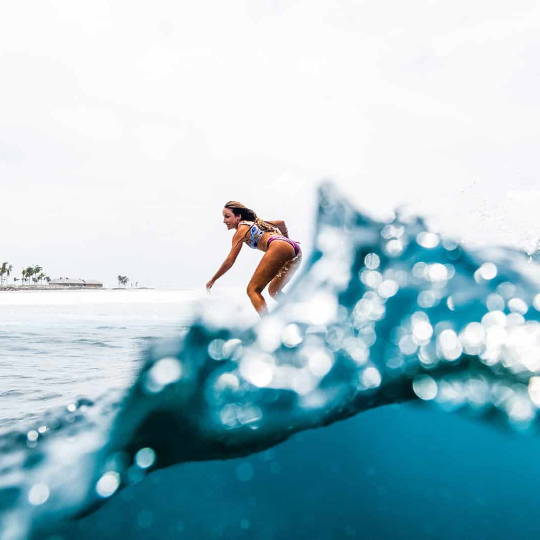 Eine Frau in einem farbenfrohen Badeanzug surft auf einer blauen Welle, aufgenommen aus einem niedrigen Winkel hinter plätscherndem Wasser, mit einem hellen Himmel und einer entfernten Küstenlinie im Hintergrund.
