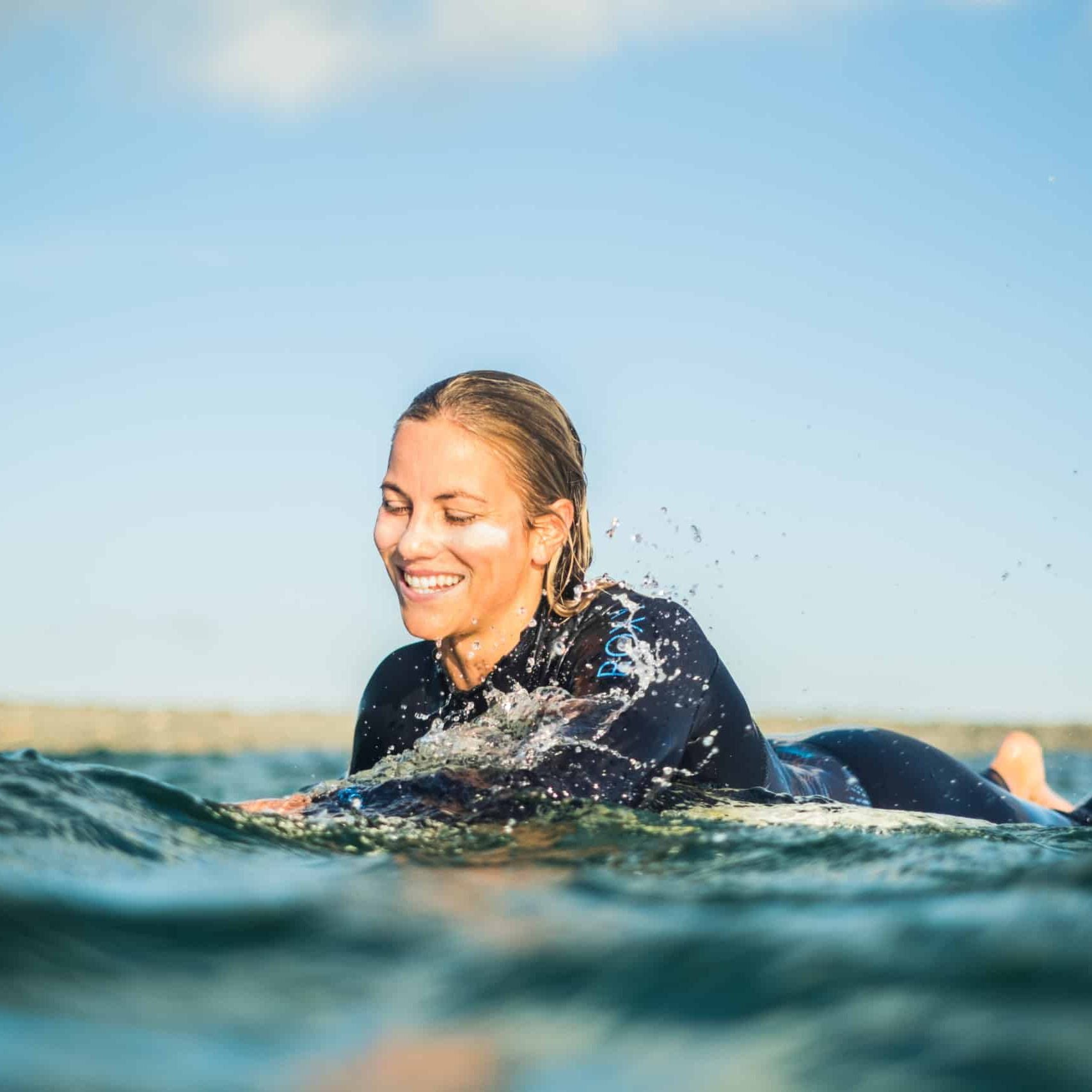Eine Frau in einem Neoprenanzug lächelt, während sie auf einem Surfbrett im Meer liegt, während das Wasser um sie herum plätschert und der blaue Himmel im Hintergrund zu sehen ist.