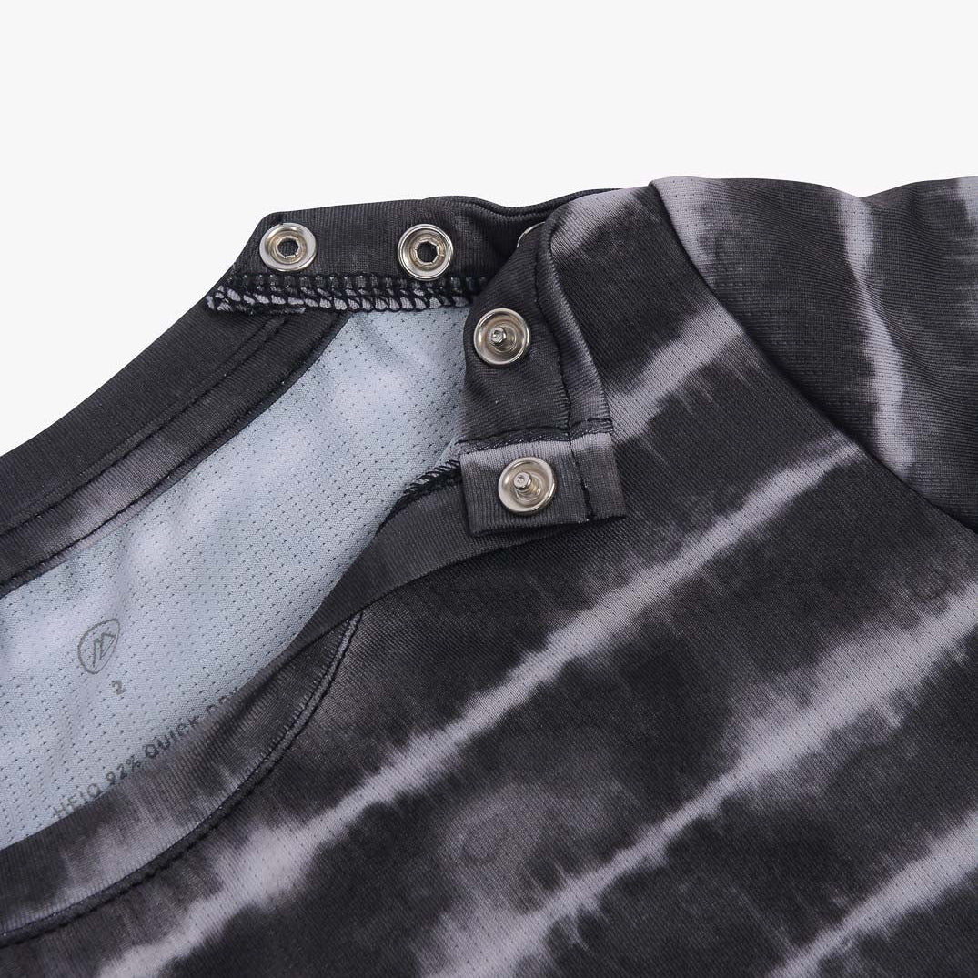 Nahaufnahme eines schwarz-grauen Batik-UV-Shirts von Aina für Kinder aus nachhaltigem Stoff mit einzigartigem Ausschnittdesign mit Druckknöpfen von Main Design. Die Druckknöpfe sind offen dargestellt und geben den Blick auf die Innenseite des Shirts frei. Der Stoff hat eine weiche Textur und das Batikmuster ist horizontal.