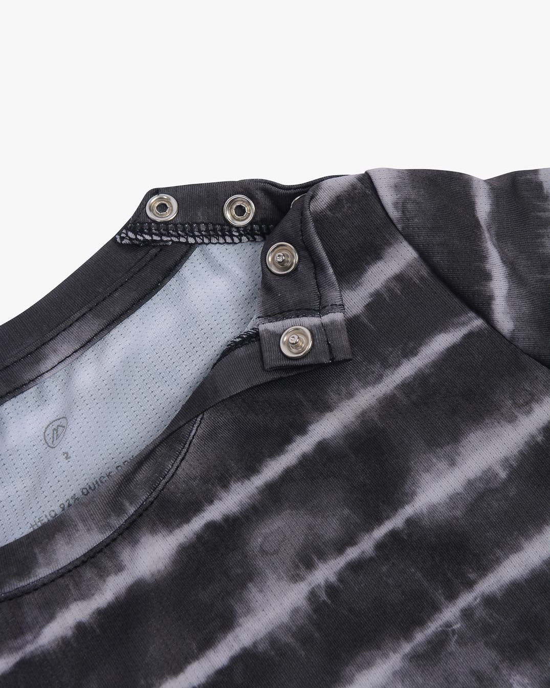 Nahaufnahme eines schwarz-grauen Batik-UV-Shirts von Aina für Kinder aus nachhaltigem Stoff mit einzigartigem Ausschnittdesign mit Druckknöpfen von Main Design. Die Druckknöpfe sind offen dargestellt und geben den Blick auf die Innenseite des Shirts frei. Der Stoff hat eine weiche Textur und das Batikmuster ist horizontal.