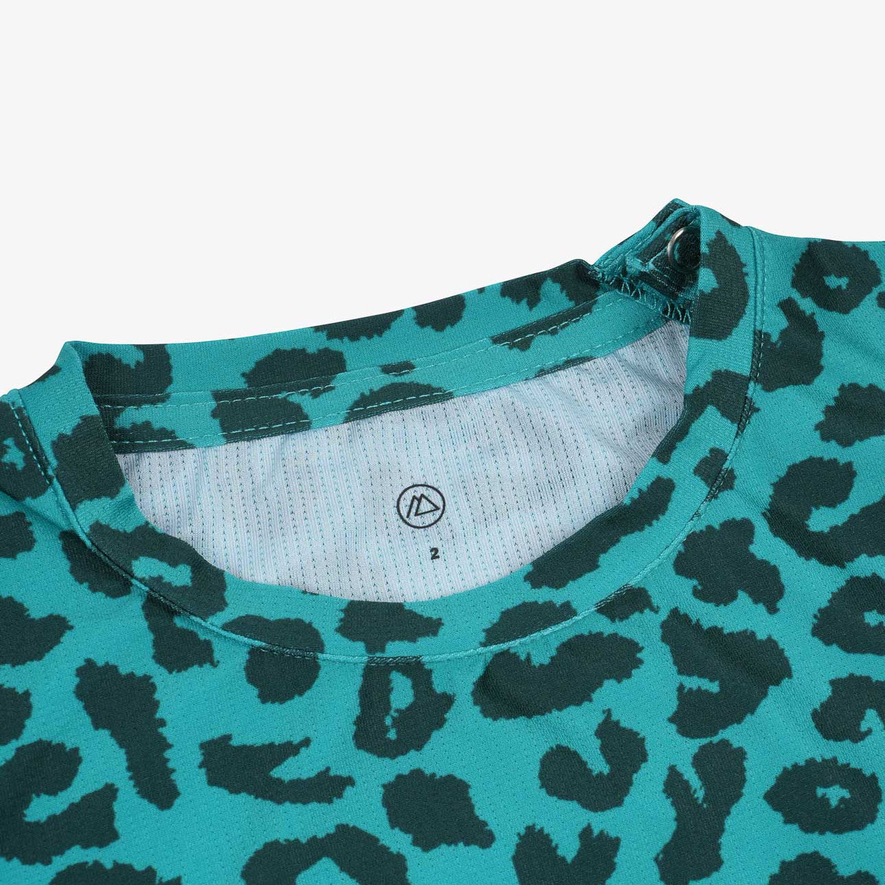 Nahaufnahme eines Aina Kinder UV-Shirts von Main Design in Blaugrün mit schwarzem Leopardenmuster. Es verfügt über einen Rundkragen und ein kleines Größenetikett. Das dehnbare, bequeme und schnelltrocknende Material bietet LSF 50+ Schutz für sonnige Abenteuer.