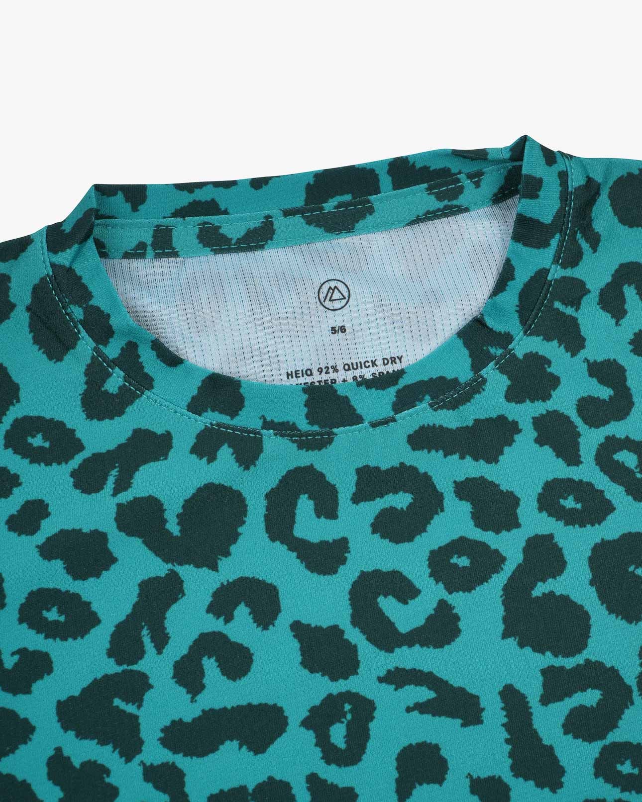 Eine Nahaufnahme des Aina Kinder UV-Shirts von Main Design in Blaugrün mit schwarzem Leopardenmuster. Ideal für sonnige Tage, bietet es LSF 50+ Schutz. Kragen und ein Teil der Brust sind sichtbar, ebenso wie ein weißes Etikett mit der Größe „S/M“ und der schnelltrocknenden Funktion.