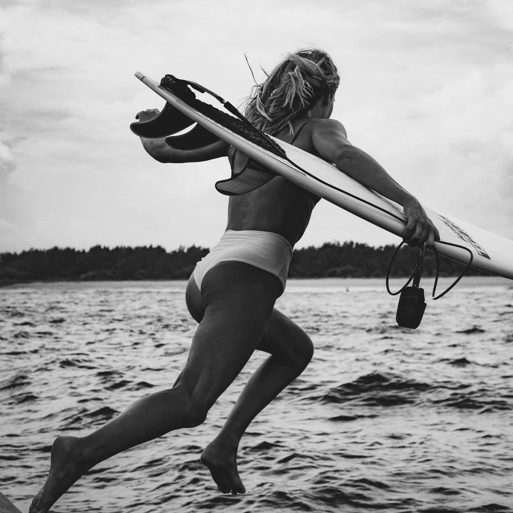 Eine Person im Badeanzug wird mitten in der Luft aufgenommen, wie sie in den Ozean läuft, während sie ein Surfbrett unter einem Arm hält. Das Bild ist in Schwarz-Weiß gehalten, mit Wasser und entfernten Bäumen im Hintergrund.
