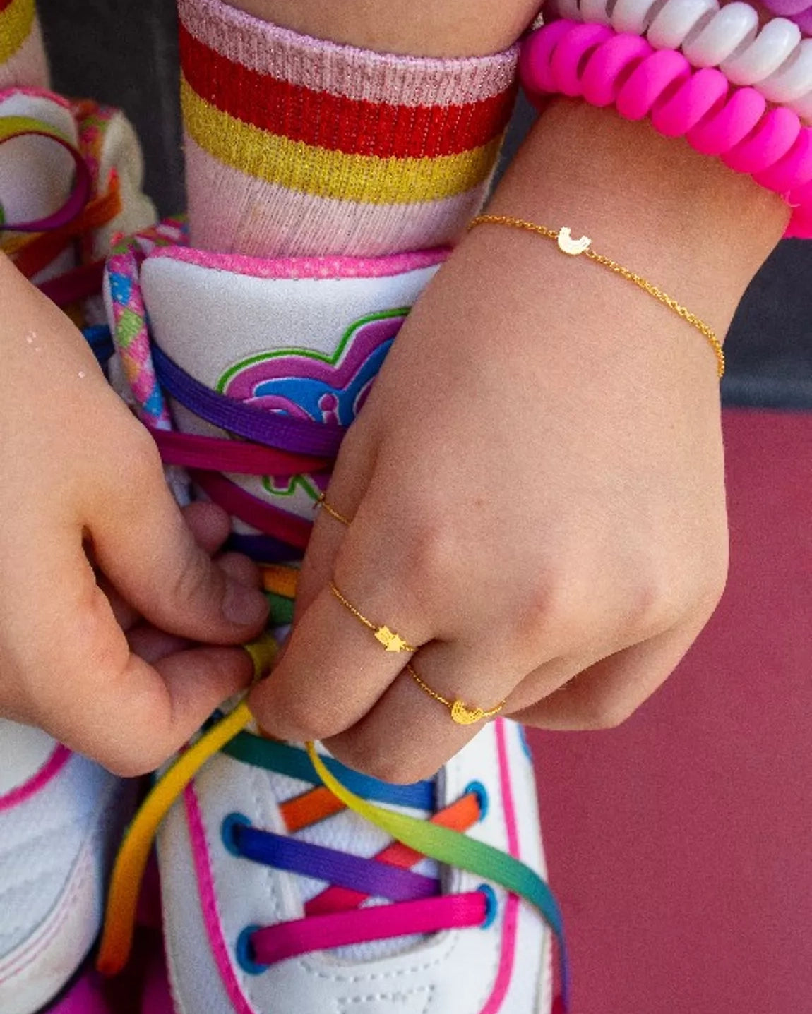 Ein Kind bindet regenbogenfarbene Schnürsenkel an weiße Turnschuhe, trägt bunt gestreifte Socken, leuchtend rosa Perlenarmbänder und ein zartes handgefertigtes Armband von Adorabili by Adorabili.