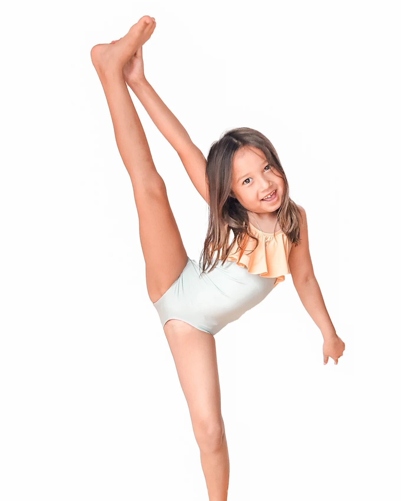 Ein junges Mädchen im Ava Mädchen Badeanzug von Main Design aus nachhaltigen Materialien führt vor einem weißen Hintergrund einen hohen, gestreckten Beinschlag aus. Mit langen Haaren und einem Lächeln zeigt sie ihre Flexibilität und Begeisterung, während die verstellbaren Träger für eine perfekte Passform sorgen.