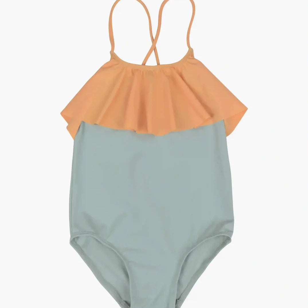 Der Ava Mädchen Badeanzug von Main Design ist ein Kinderbadeanzug mit einem hellblauen Unterteil und einem pfirsichfarbenen Rüschenoberteil. Er hat dünne, gekreuzte Träger auf der Rückseite, was ihn schlicht und zum Schwimmen geeignet macht. Der Hintergrund ist schlicht weiß.