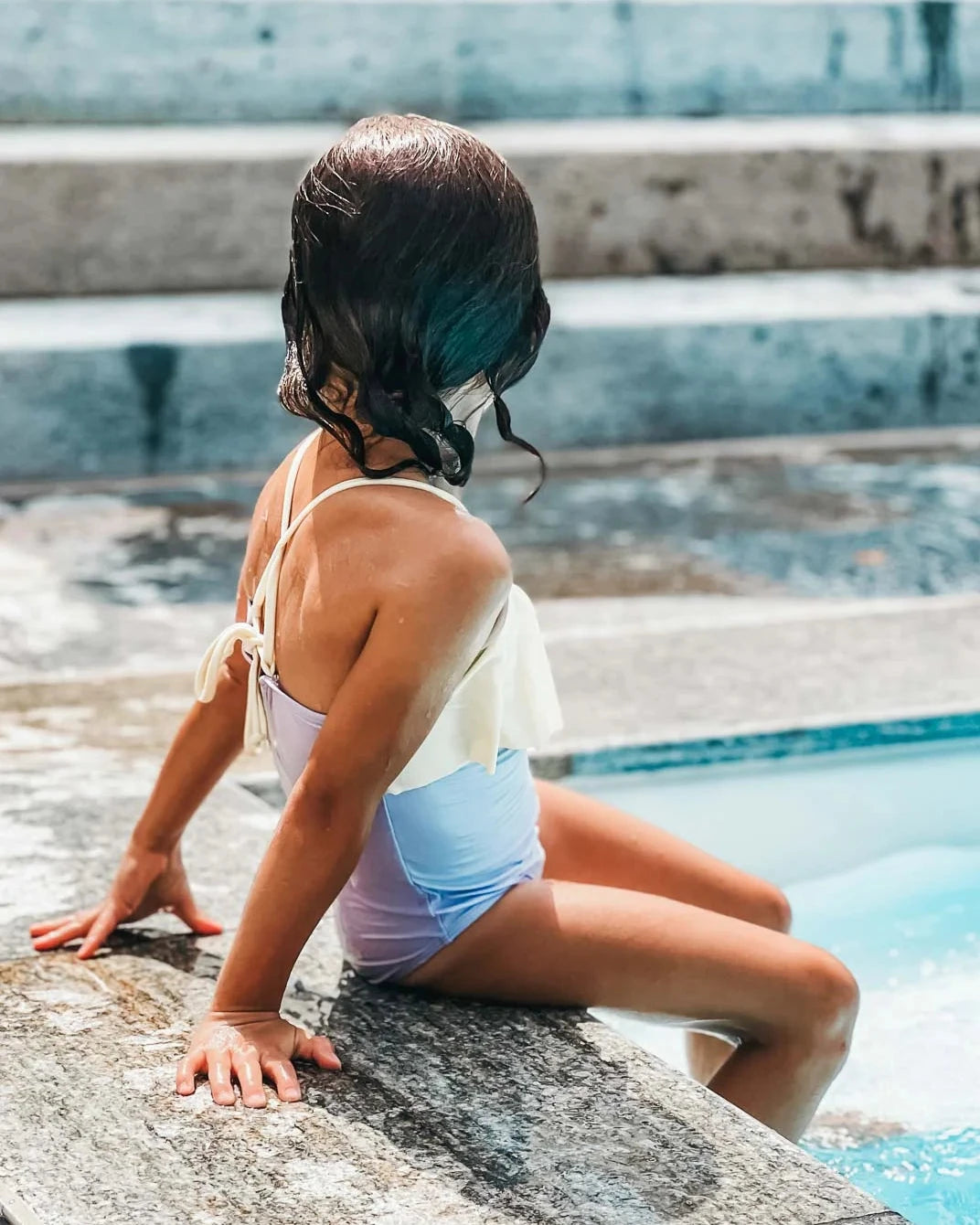 Ein kleines Kind sitzt am Rand eines Pools und blickt von der Kamera weg. Das Kind trägt einen hellen Ava Mädchen Badeanzug von Main Design mit verstellbaren Trägern und hat nasse Haare. Es legt seine Hand auf den Poolrand, im Hintergrund sind Stufen zu sehen. Die Szene ist hell und sonnig und lässt auf einen warmen Sommertag schließen.