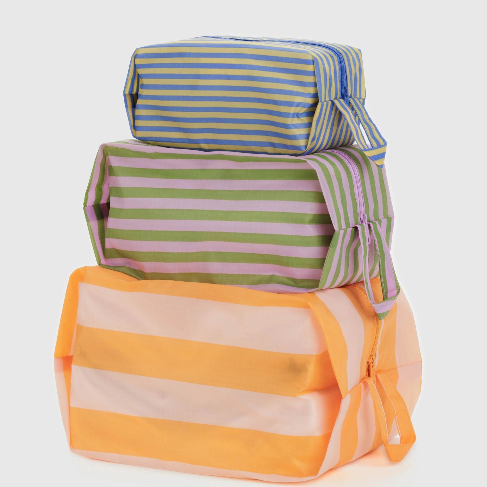 Drei rechteckige BAGGU 3D Zip Set Taschen der Marke BAGGU mit blauen, grünen und orangen Streifen sind als stilvolle Reise-Organizer gestapelt und vor weißem Hintergrund arrangiert.