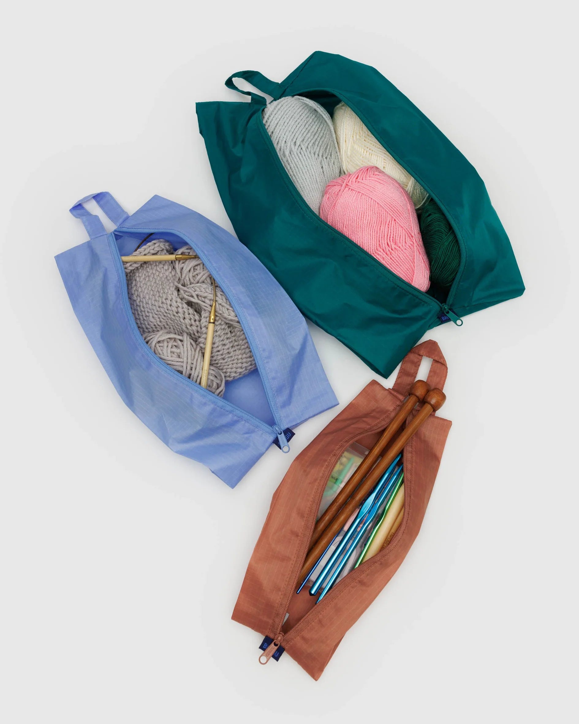 Das BAGGU 3D Zip Set von BAGGU besteht aus blauen, grünen und braunen Reißverschlusstaschen für Strickutensilien auf einer weißen Oberfläche - ideal als umweltfreundliche Taschen oder praktische Reisebegleiter.