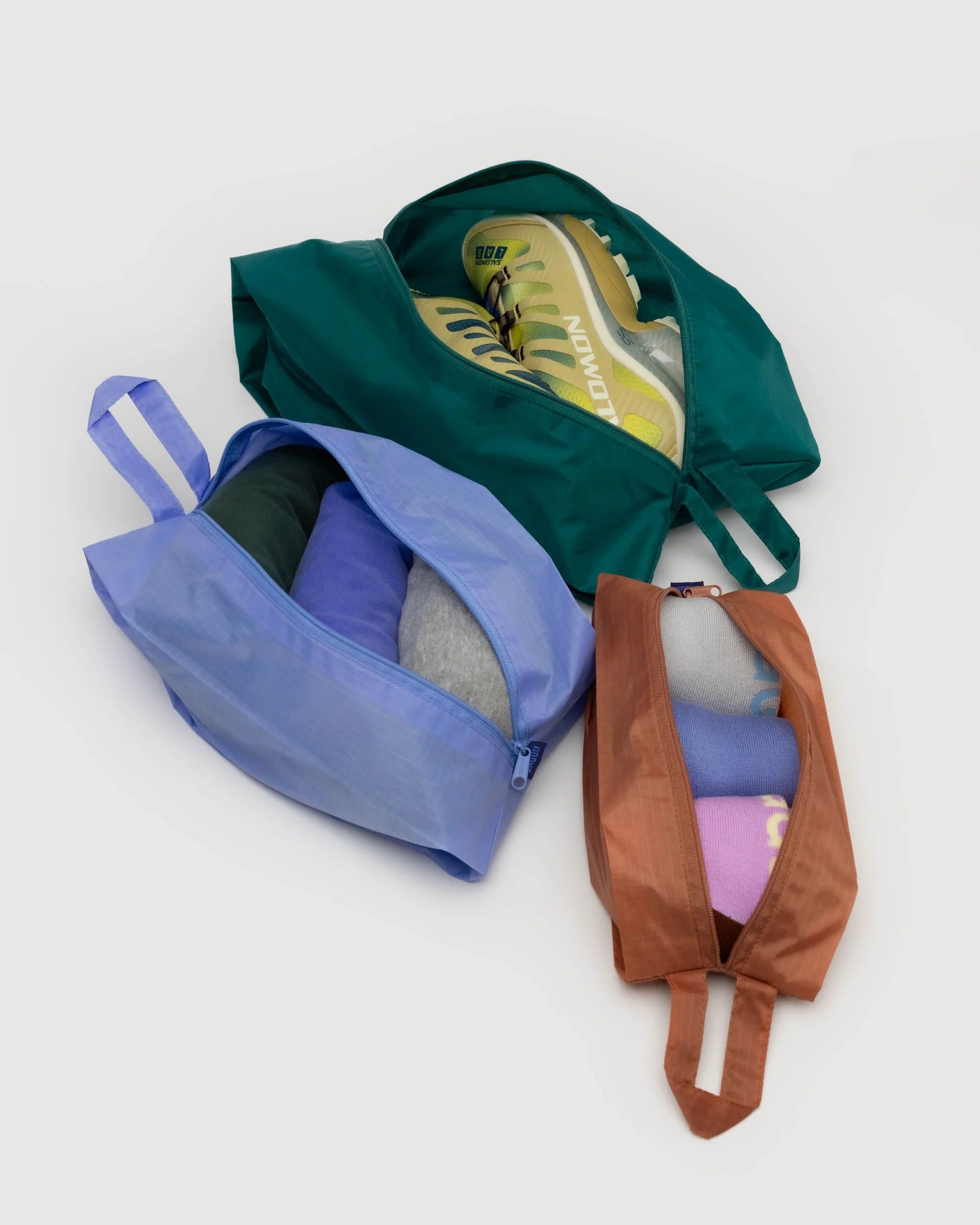 Abgebildet ist das BAGGU 3D Zip Set von BAGGU in grün, blau und braun. Im grünen Würfel werden Turnschuhe aufbewahrt, in den blauen und braunen Würfeln gefaltete Kleidung. Alle Organizerwürfel haben robuste Griffschlaufen aus Stoff zum einfachen Tragen.