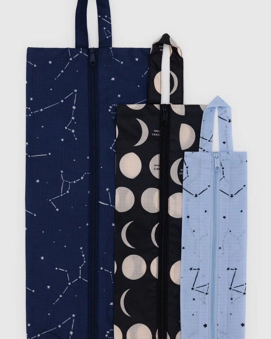 Das BAGGU 3D Zip Set von BAGGU enthält drei stylische, nachhaltige Reißverschlusstaschen: navy mit weißen Sternen, schwarz mit cremefarbenen Mondphasen, und hellblau mit dunklen Konstellationen - perfekt als Reise-Organizer nebeneinander angeordnet.