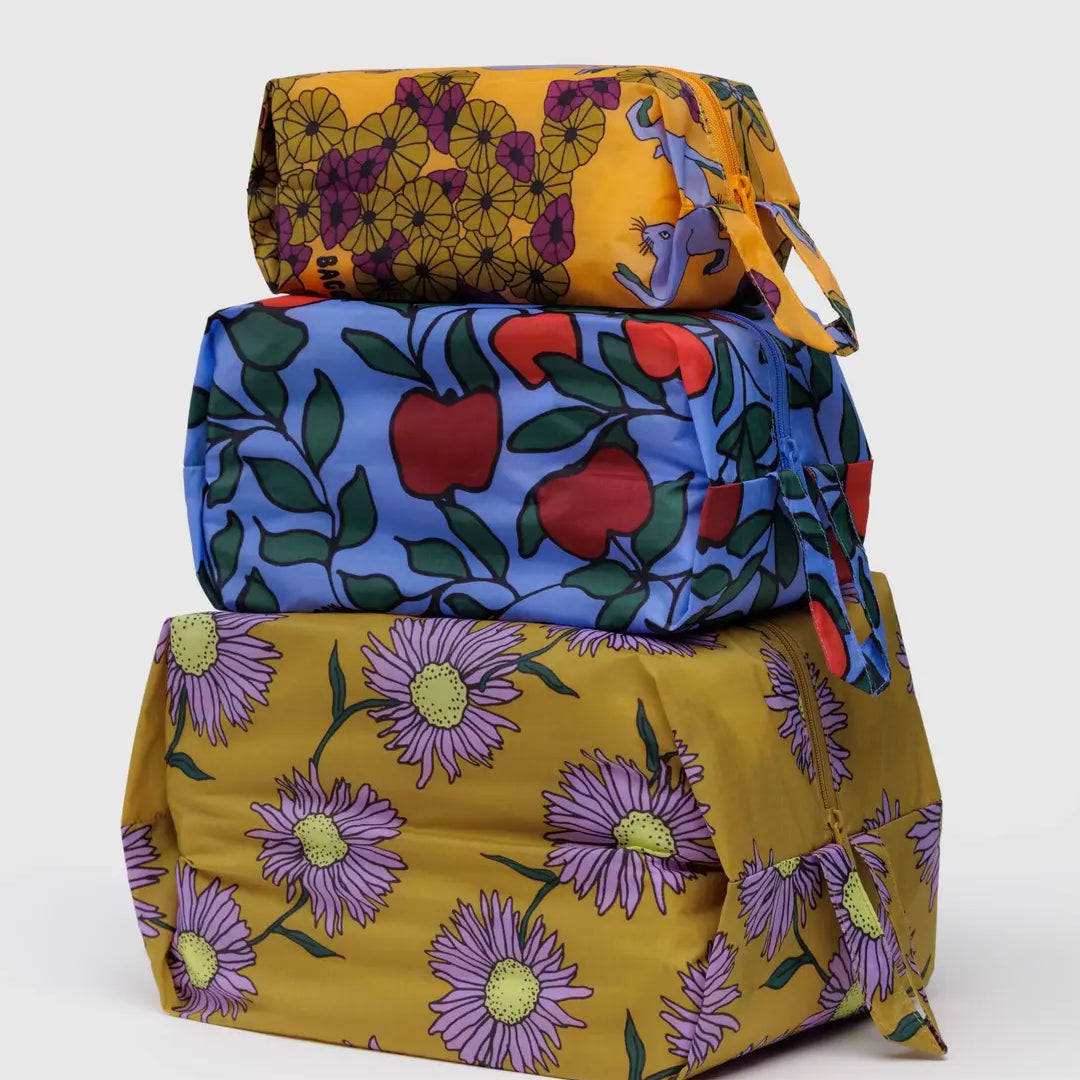 Drei in Stoff eingewickelte Pakete - ein gelbes mit lila Blume, ein blaues mit rotem Apfel und ein gelb-grünes mit lila Gänseblümchen - sind gestapelt, ideal zum Organisieren im BAGGU 3D Zip Set von BAGGU.