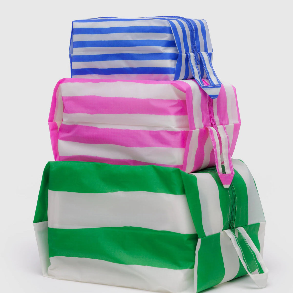 Das BAGGU 3D Zip Set von BAGGU besteht aus drei rechteckigen Reißverschlusstaschen mit blauen, pinken und grünen Streifen, die gestapelt als stilvolle Reise-Organizer präsentiert werden.
