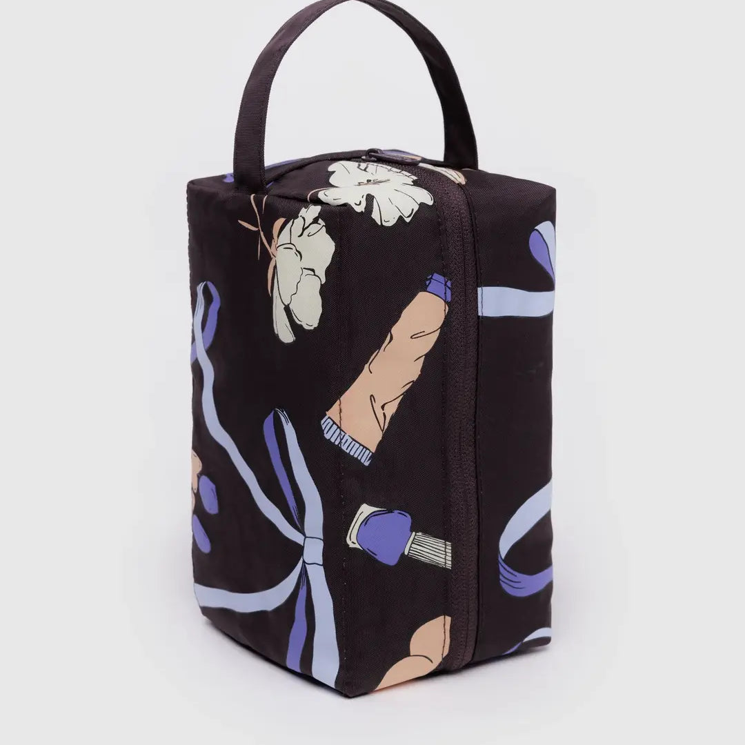 Das BAGGU Dopp Kit Necessaire von BAGGU ist ein schwarzer, rechteckiger Kulturbeutel aus recyceltem Nylon, mit einem Griff an der Oberseite und einem bunten blauen Band und beigen Händen, die aufrecht auf einem weißen Hintergrund stehen.