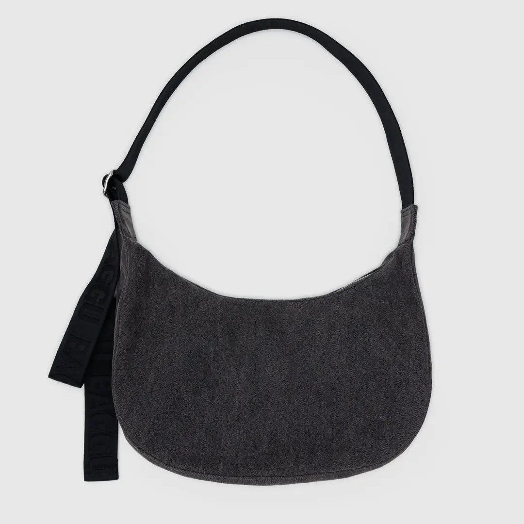 Die BAGGU Medium Denim Crescent Bag von BAGGU ist eine dunkelgraue, halbmondförmige Tasche mit einem breiten schwarzen Riemen und einem schwarzen Stoffanhänger auf der linken Seite. Sie ist aus recycelter Baumwolle gefertigt und auf einem einfarbigen hellgrauen Hintergrund abgebildet.