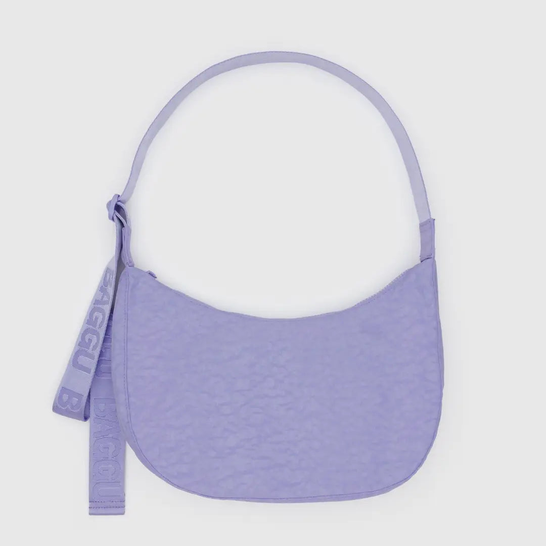 Eine lavendelfarbene BAGGU Medium Nylon Crescent Bag mit geschwungener Silhouette, passendem Gurt, strukturierter Oberfläche und einem auffälligen "BAGGU"-Anhänger wird vor einem schlichten weißen Hintergrund präsentiert.