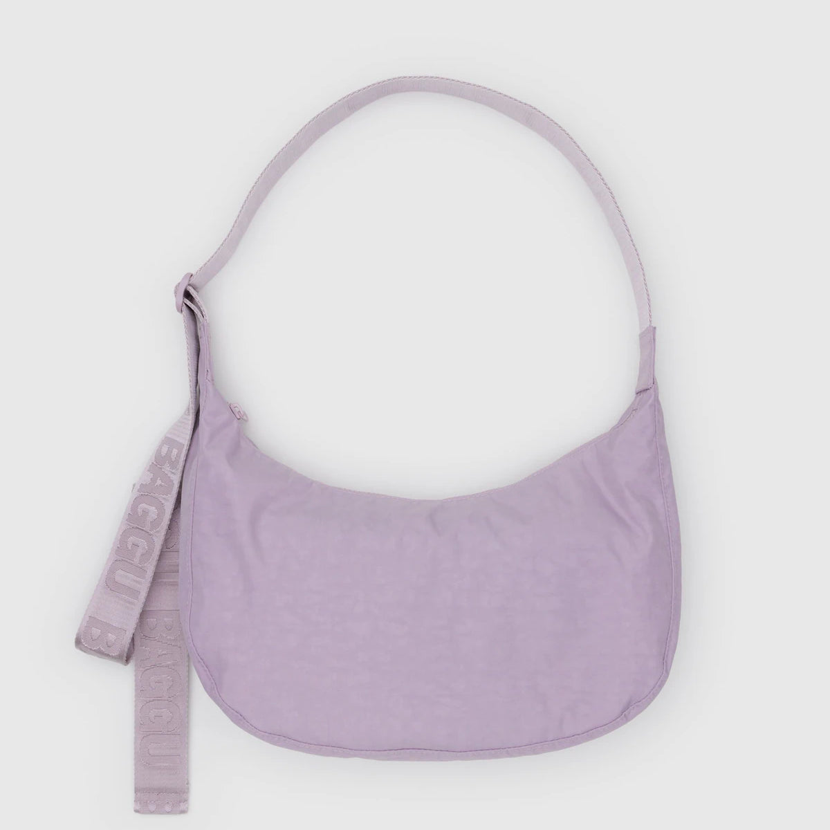 Die BAGGU Medium Nylon Crescent Bag in pastellfarbenem Lavendel hat eine geschwungene Form, einen verstellbaren Riemen und einen passenden abnehmbaren "BAGGU"-Schlüsselanhänger auf der linken Seite, der auf einem hellgrauen Hintergrund abgebildet ist.