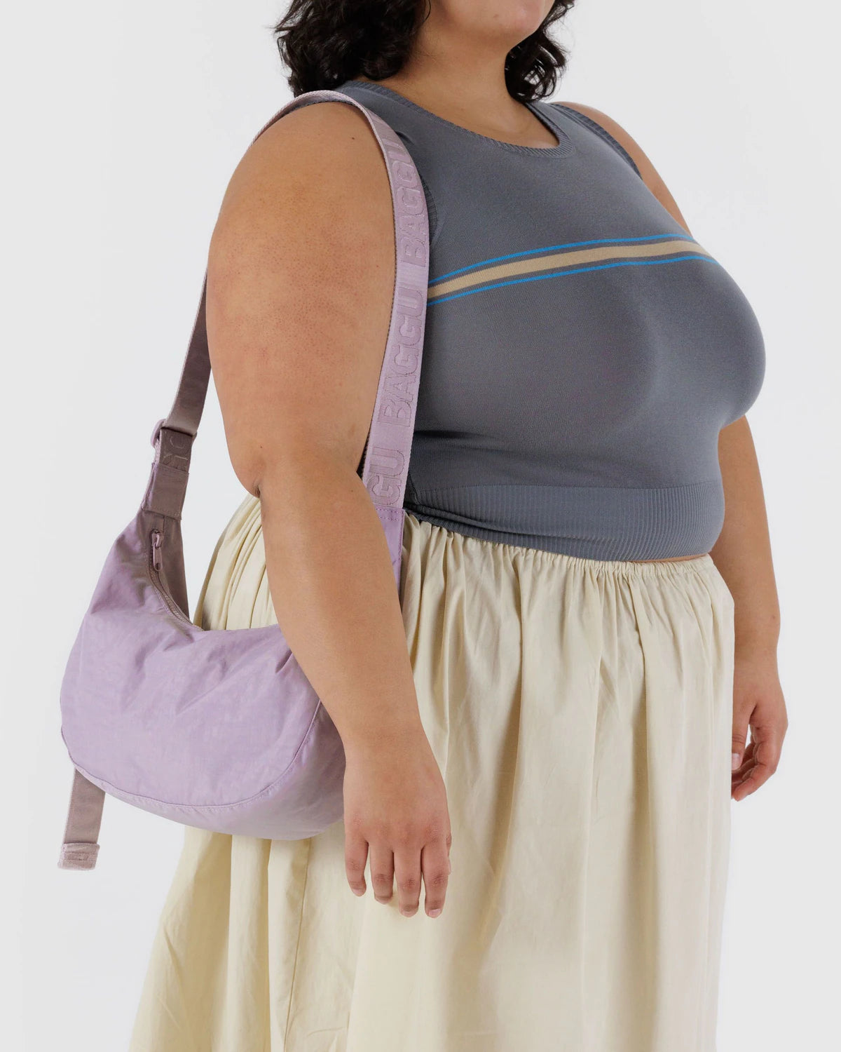 Eine Person, die ein ärmelloses graues Oberteil mit blauen Streifen und einen cremefarbenen Rock trägt und eine BAGGU Medium Nylon Crescent Bag von BAGGU mit sich führt, steht vor einem schlichten weißen Hintergrund.