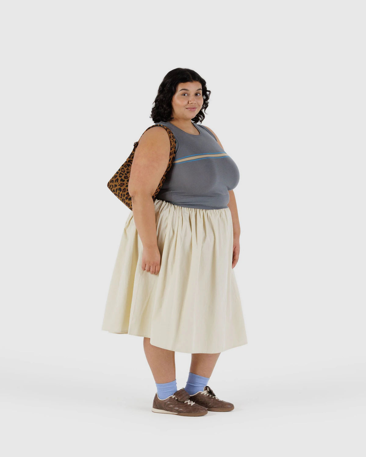 Eine Frau mit kurzen lockigen dunklen Haaren trägt ein graues ärmelloses Oberteil, einen cremefarbenen Rock, blaue Socken, braune Schuhe und trägt eine BAGGU Mini-Nylon-Schultertasche mit Leopardenmuster, während sie vor einem schlichten weißen Hintergrund steht.