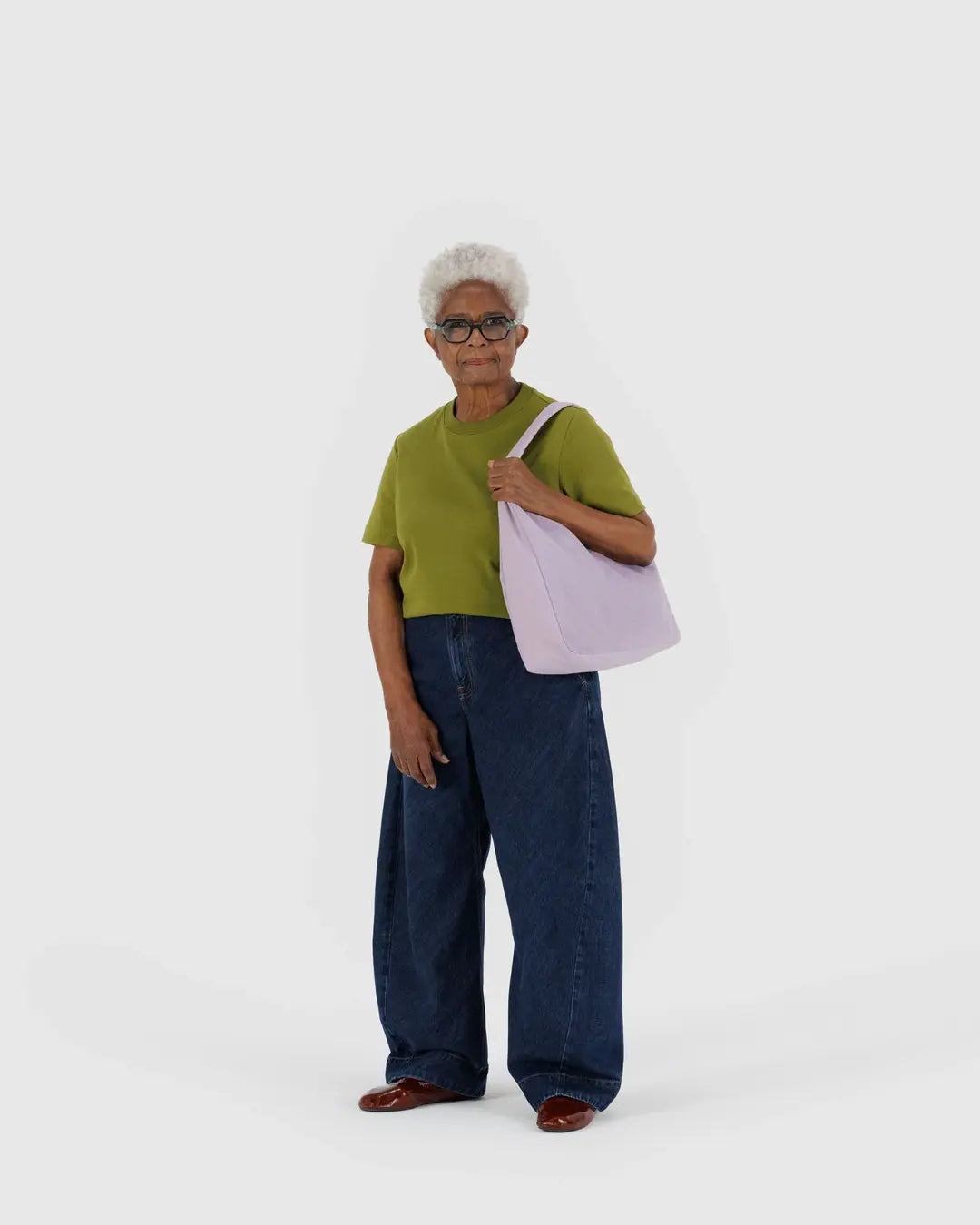 Eine ältere Frau mit kurzen weißen Haaren trägt ein grünes Hemd, weite Bluejeans, braune Schuhe und trägt eine BAGGU Nylon Shoulder Bag von BAGGU über der Schulter. Sie steht vor einem schlichten weißen Hintergrund.