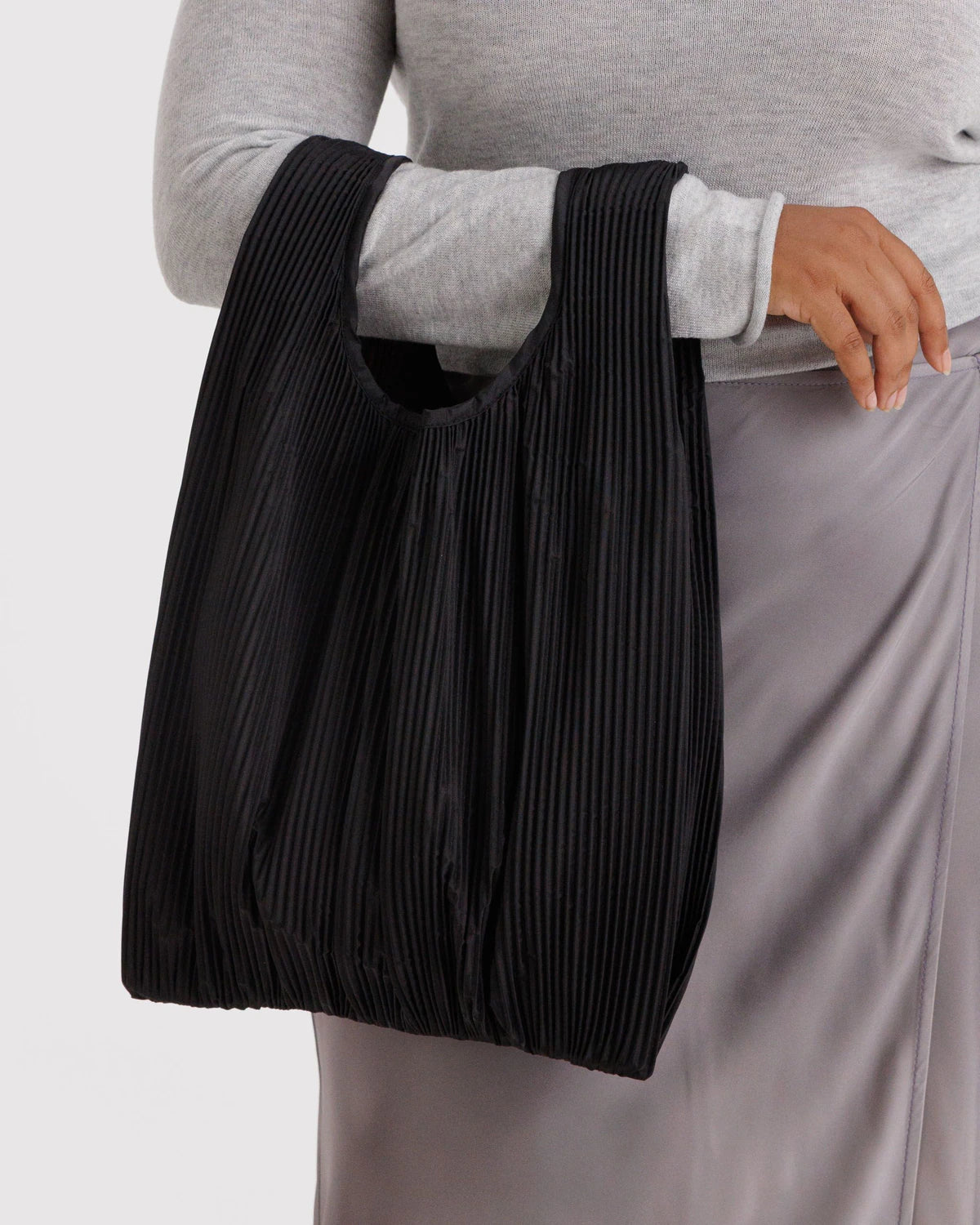 Eine Person in einem hellgrauen, langärmeligen Oberteil und einem seidengrauen Rock hält die BAGGU Pleated Tragetasche, eine schwarze, plissierte Allzwecktasche mit runden Henkeln, die aus recyceltem Ripstop-Nylon von BAGGU hergestellt wird.