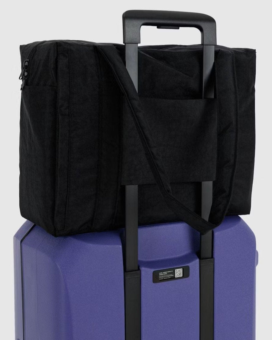 Eine BAGGU Small Cloud Carry-On Tasche aus schwarzem, recyceltem Nylon ist auf einem lilafarbenen Hartschalenkoffer mit verlängertem Griff befestigt, dessen Gurte über den Koffer drapiert sind - perfekt als BAGGU Untersitz-Reisetasche.
