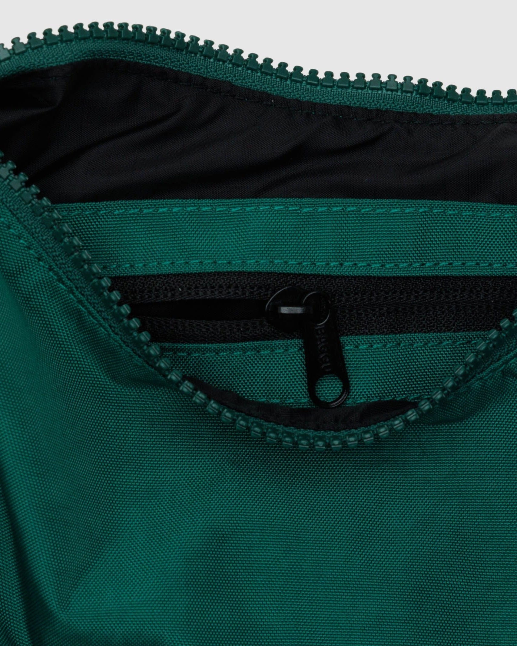 Nahaufnahme einer grünen BAGGU Small Nylon Crescent Bag, die teilweise geöffnet wurde, um eine kleinere Innentasche mit schwarzem Reißverschluss zu zeigen. Die Tasche aus recyceltem Nylon von BAGGU ist robust, wasserabweisend und perfekt für den umweltbewussten Gebrauch unterwegs.