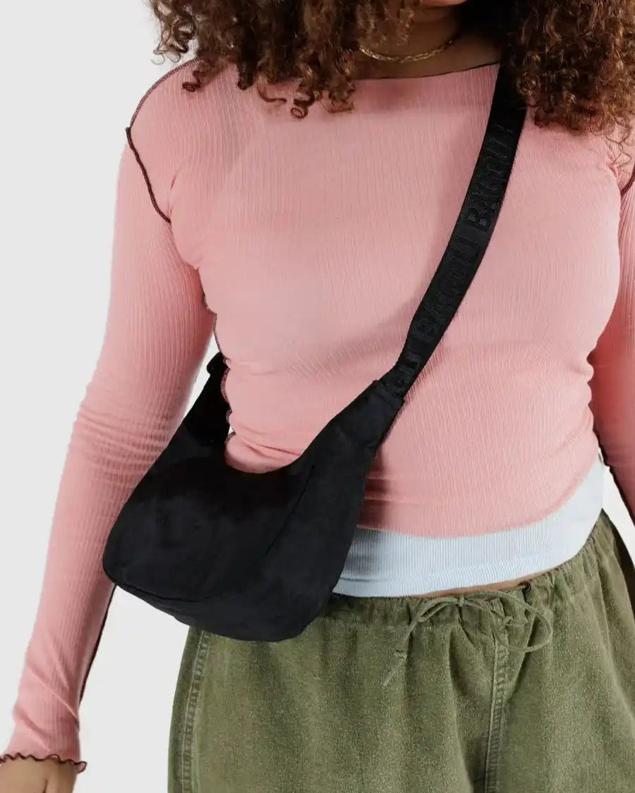 Eine Person trägt ein hellrosa langärmeliges Oberteil über einem weißen Hemd, eine olivgrüne Hose und trägt die BAGGU Small Nylon Crescent Bag in Schwarz. Das Foto ist an den Schultern und Knien abgeschnitten.