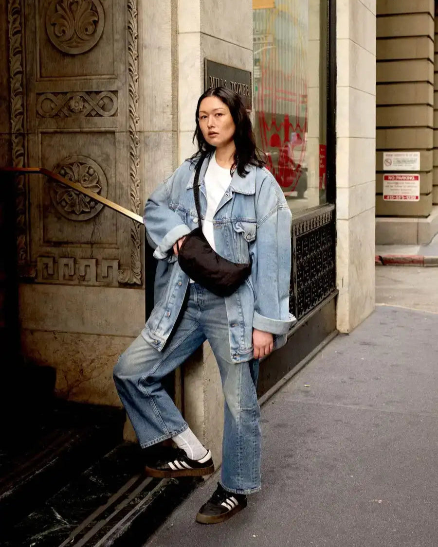 Eine Person steht auf einem städtischen Bürgersteig vor einem Gebäude, trägt eine hellblaue Oversized-Jeansjacke und Jeans, ein weißes T-Shirt, schwarze Adidas-Turnschuhe und trägt die BAGGU Small Nylon Crescent Bag.