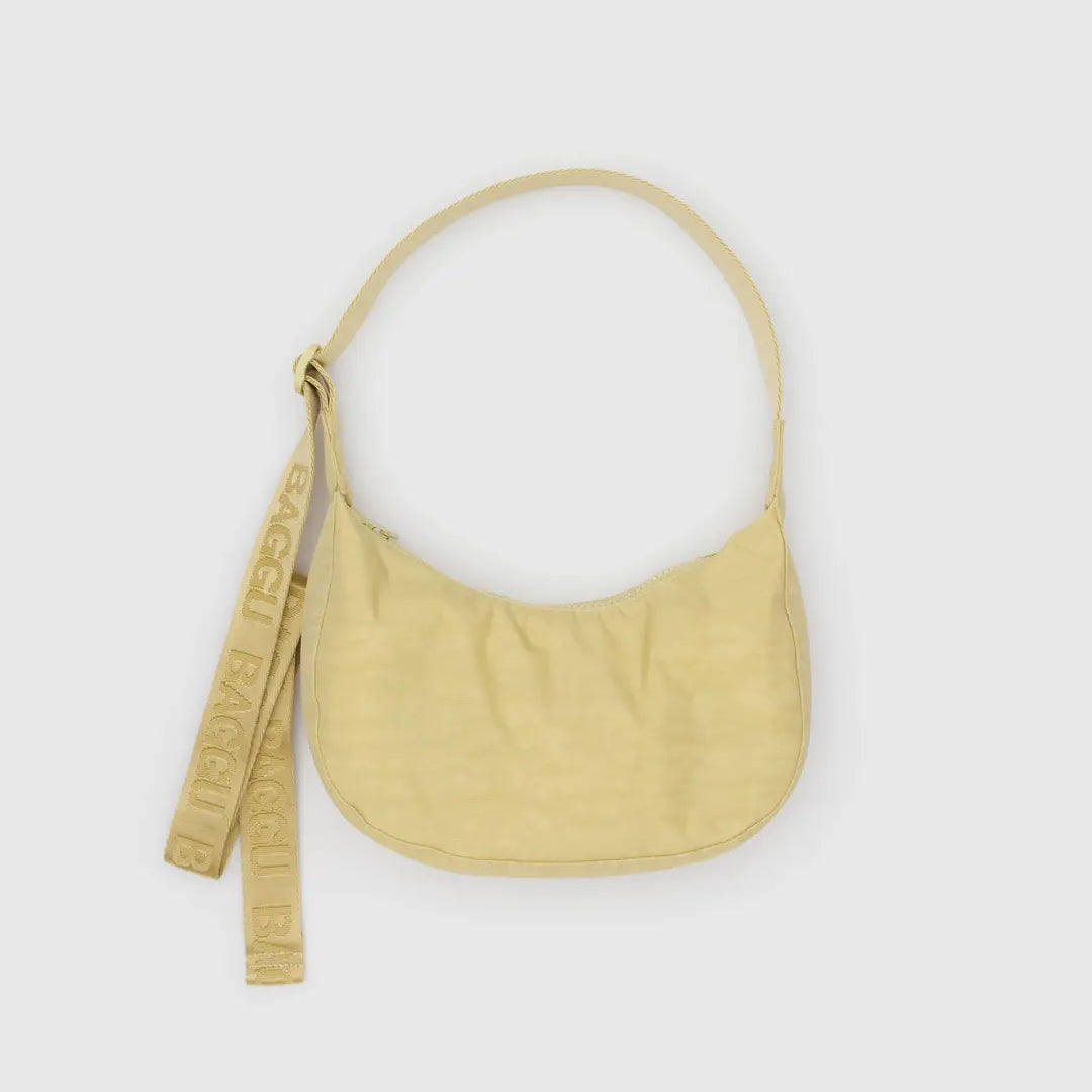 Die BAGGU Small Nylon Crescent Bag aus hellgelbem, recyceltem Nylon zeichnet sich durch eine geschwungene Silhouette, einen kurzen Riemen und zwei angebrachte Anhänger mit erhabenem "BAGGU"-Schriftzug vor einem schlichten, hellen Hintergrund aus.