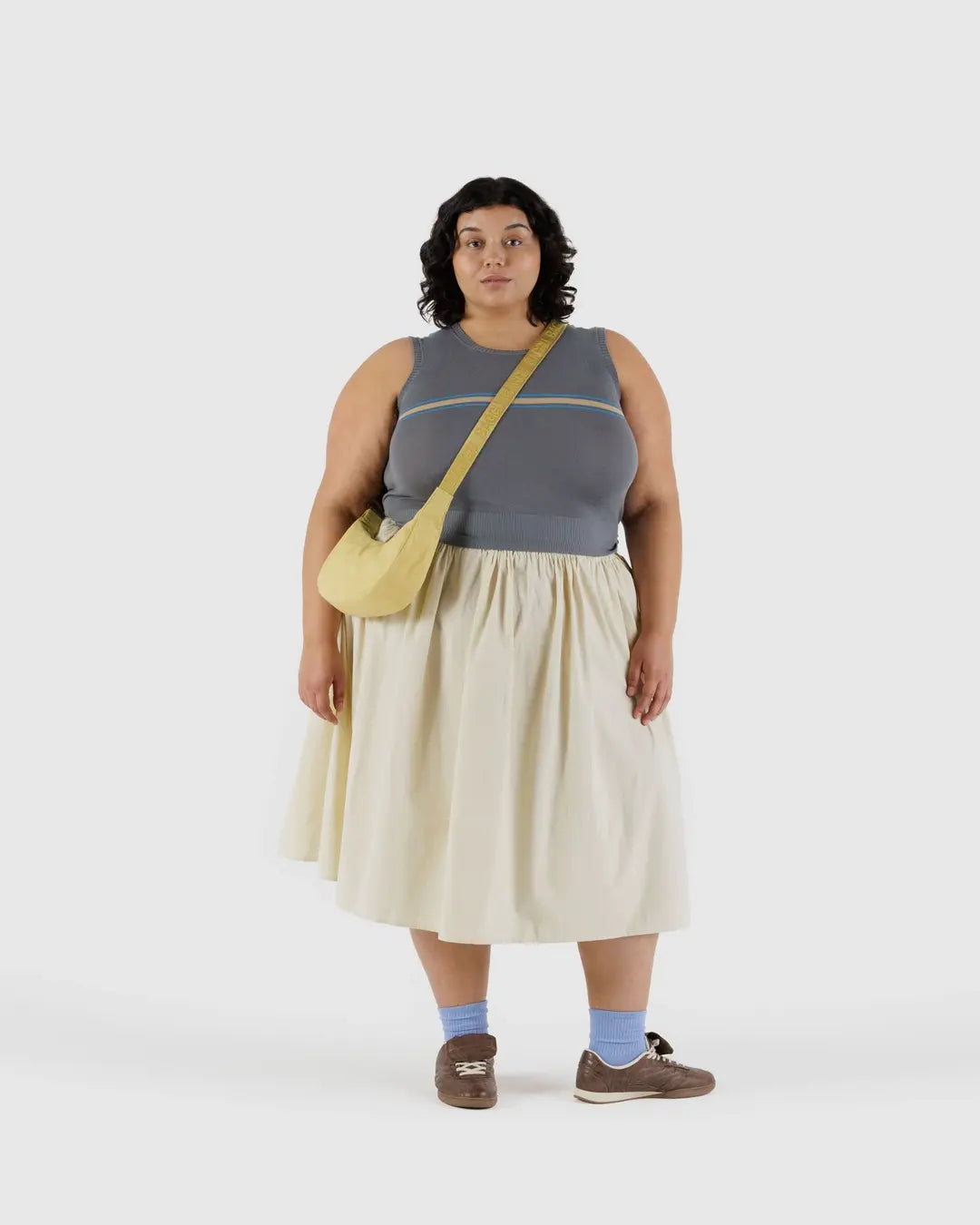 Eine Frau mit lockigem dunklem Haar trägt ein graues ärmelloses Oberteil, einen cremefarbenen Rock, braune Schuhe mit blauen Socken und trägt die BAGGU Small Nylon Crescent Bag von BAGGU vor einem schlichten hellen Hintergrund.
