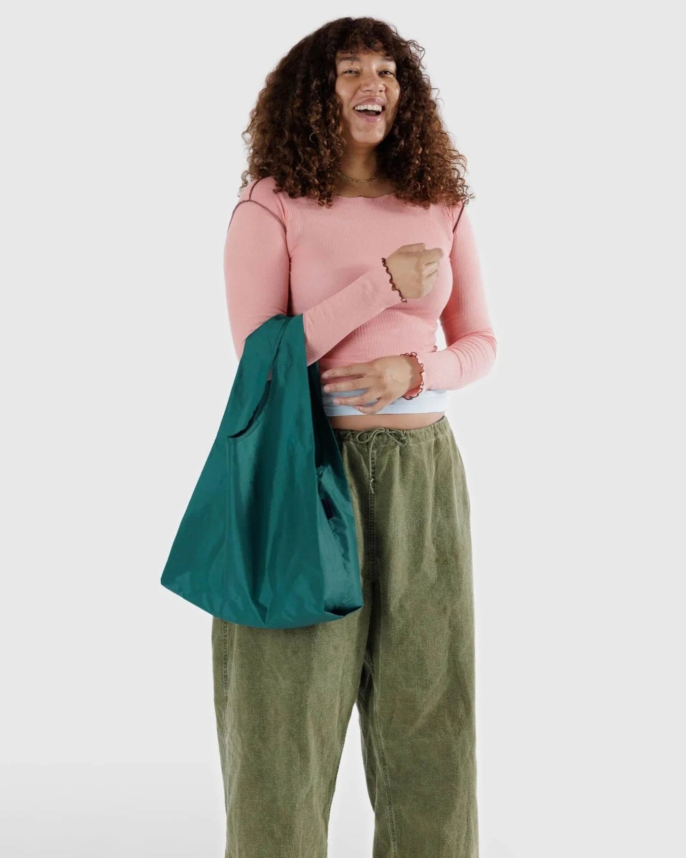 Eine Person mit lockigem Haar steht lachend da, bekleidet mit einem hellrosa Langarmshirt und einer weiten grünen Hose. Sie hält eine BAGGU Standard Tragetasche von Main Design über der linken Schulter und trägt helle Schuhe. Der Hintergrund ist schlicht hellgrau.