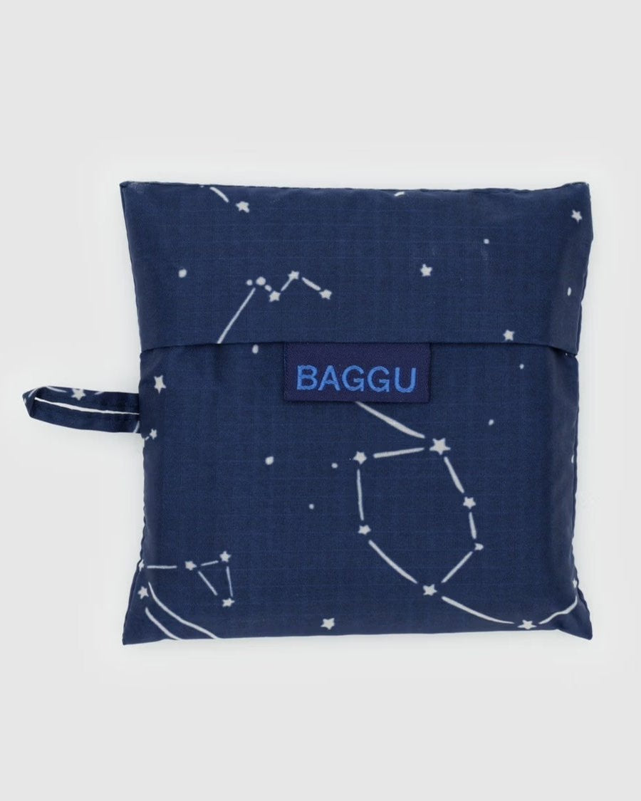 Eine BAGGU Standard Tragetasche von BAGGU in Marineblau mit Sternbildmuster, hergestellt aus recyceltem Ripstop-Nylon. Die faltbare Tasche hat einen kleinen Griff auf der linken Seite und ist vor einem schlichten, hellen Hintergrund gehalten.
