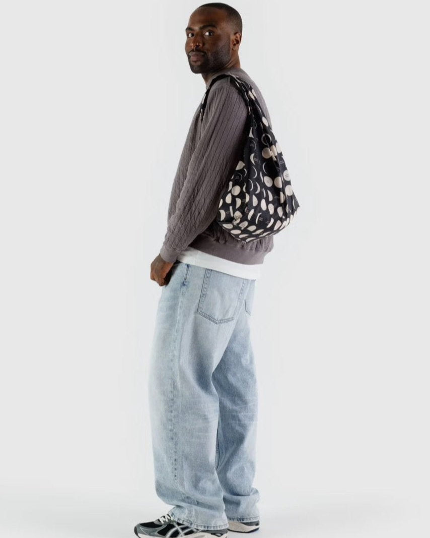 Ein Mann in grauem Strukturpullover, weißem T-Shirt, hellblauen Baggy-Jeans und grauen Turnschuhen steht seitlich mit einer BAGGU Standard Tragetasche von BAGGU aus recyceltem Ripstop-Nylon vor einem schlichten hellen Hintergrund.