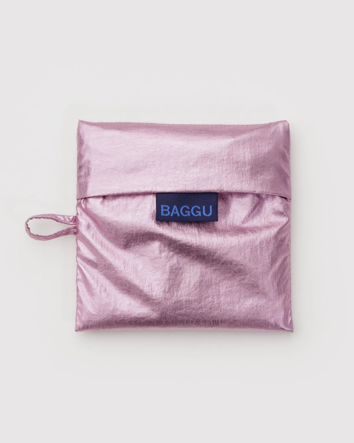 Metallicrosa, quadratische BAGGU Standard Tragetasche aus recyceltem Ripstop-Nylon, gefaltet mit Überschlag und Seitenschlaufe, mit blauem "BAGGU"-Label.