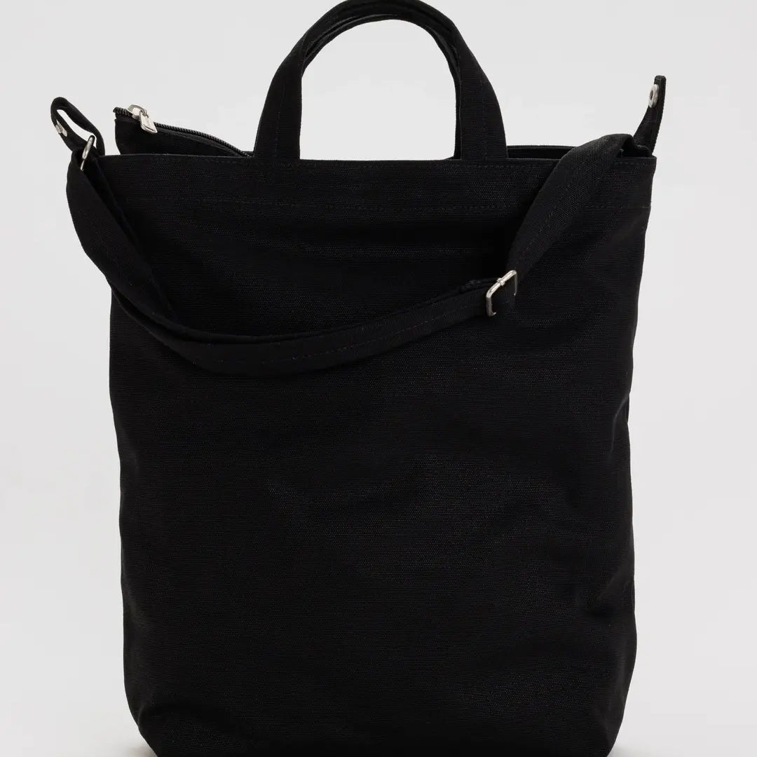 Die BAGGU Zip Duck Bag aus recycelter Baumwolle in Schwarz mit kurzen Henkeln, langem verstellbarem Schulterriemen und Reißverschluss von BAGGU vor hellgrauem Hintergrund.