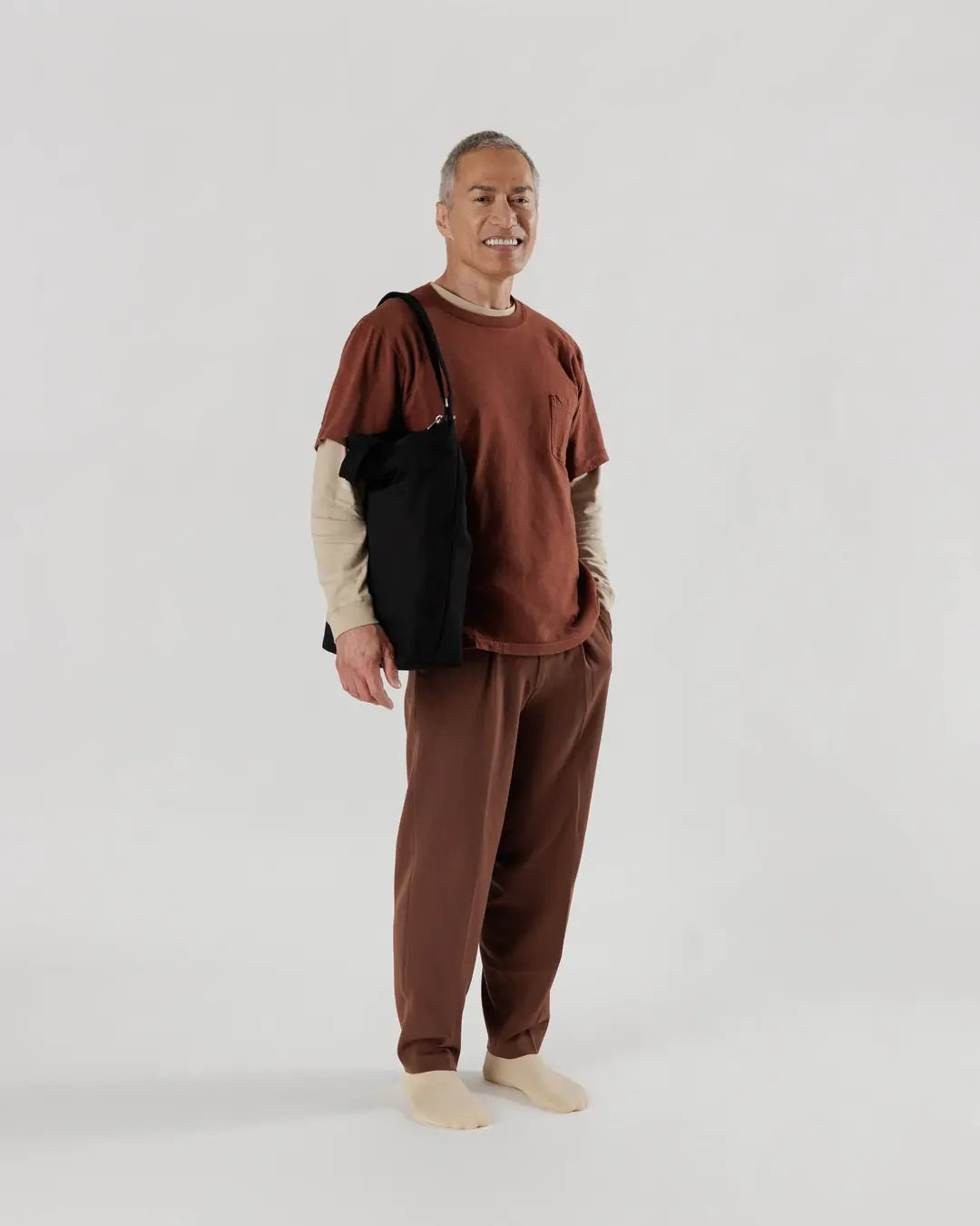 Ein älterer Mann mit kurzen grauen Haaren lächelt vor einem schlichten Hintergrund. Er trägt ein braunes T-Shirt, ein beiges langärmeliges Hemd, eine passende Hose, cremefarbene Socken und hat die BAGGU Zip Duck Bag von BAGGU aus recycelter Baumwolle dabei.