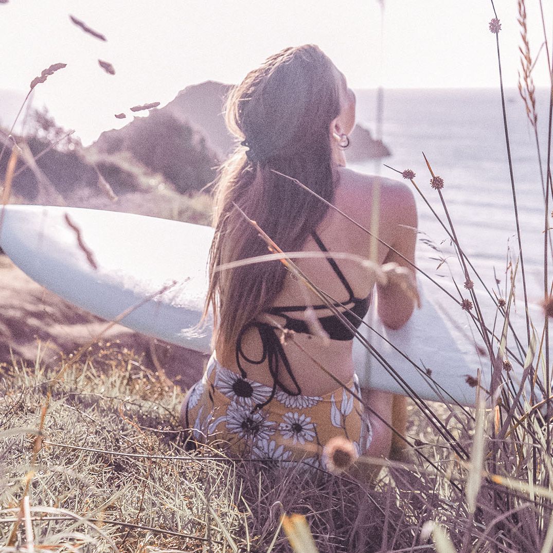 Eine Frau im Badeanzug sitzt an einem sonnigen Tag mit einem Surfbrett in der Hand auf einer Wiese in Küstennähe und blickt auf das Meer hinaus. Hohes Gras und Wildblumen umgeben sie.
