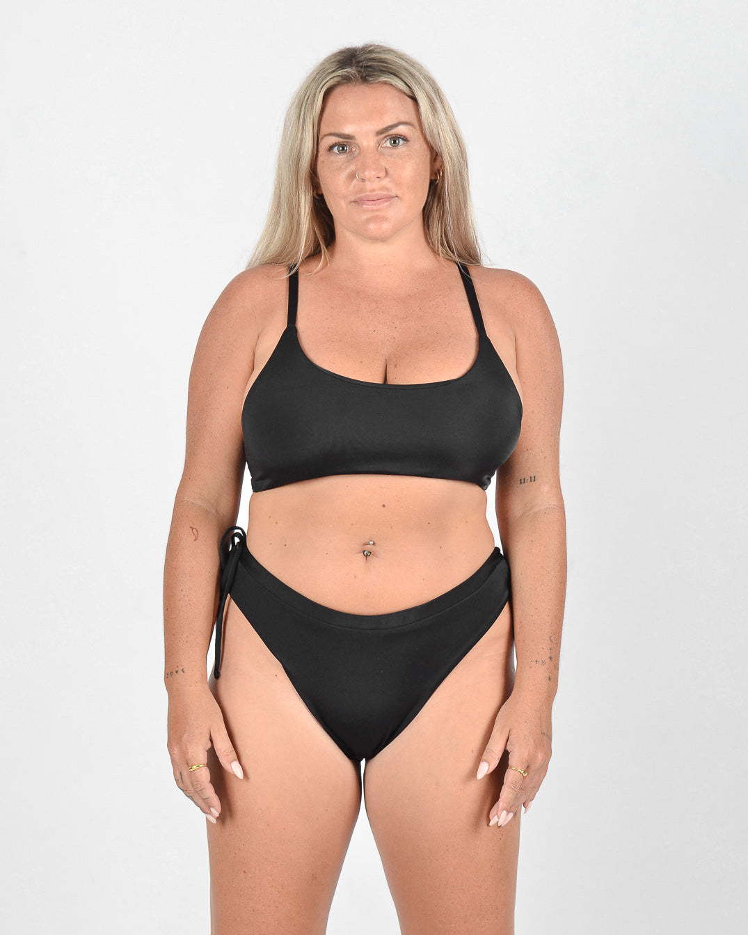 Eine Frau mit langen blonden Haaren trägt eine Blush Surf Bikinihose von Main Design aus umweltfreundlichem Stoff. Sie steht mit neutralem Gesichtsausdruck und entspannten Armen vor einem schlichten weißen Hintergrund und blickt in die Kamera.